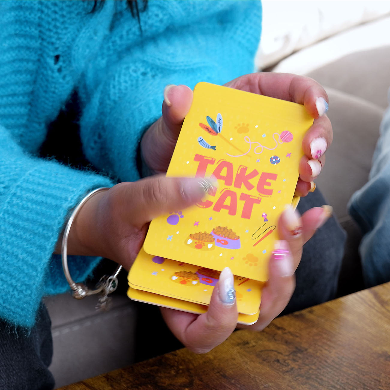 A person holding a deck of orange 'TAKE CAT' cards with colourful illustrations, wearing a bright turquoise knit sweater and a silver bangle.