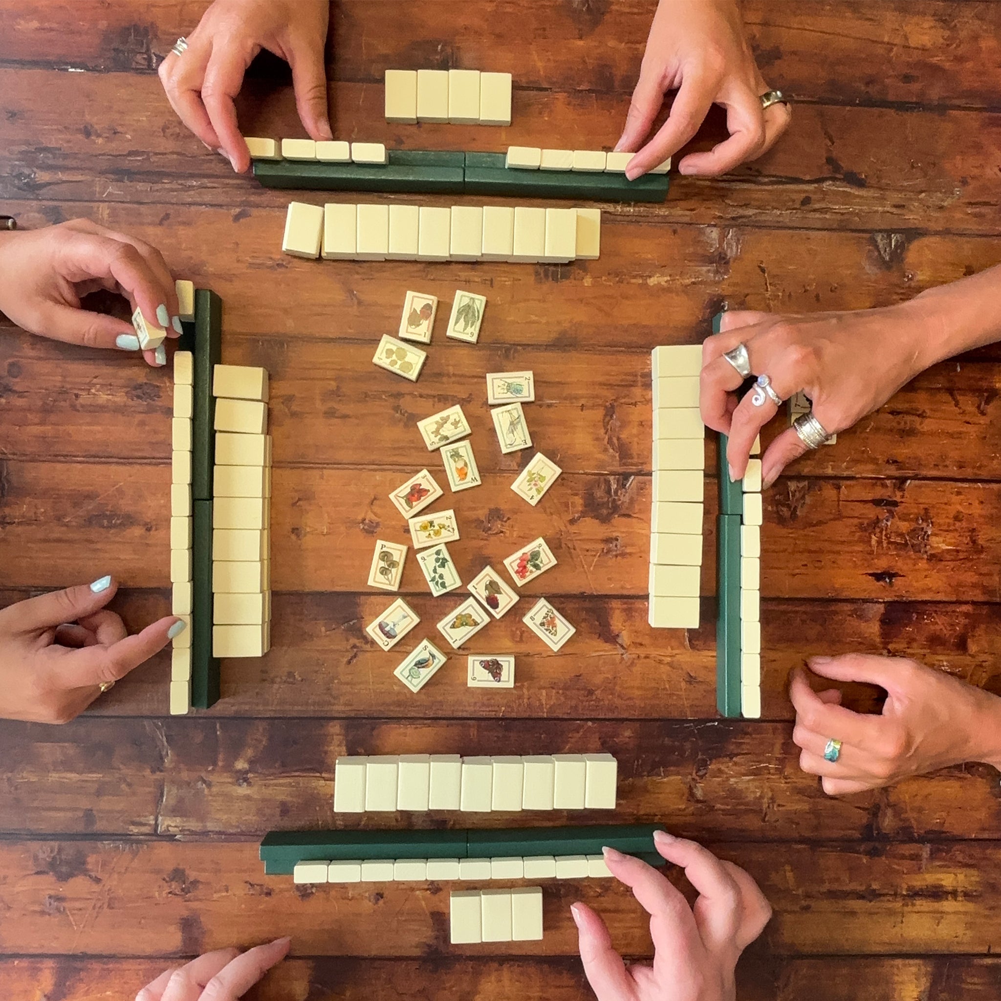 An overhead view of a game of mahjong being played by four people, whose hands and arms are visible around a wooden table. The players have arranged dark green racks of cream-coloured tiles, with several tiles scattered in the center of the wooden surface.