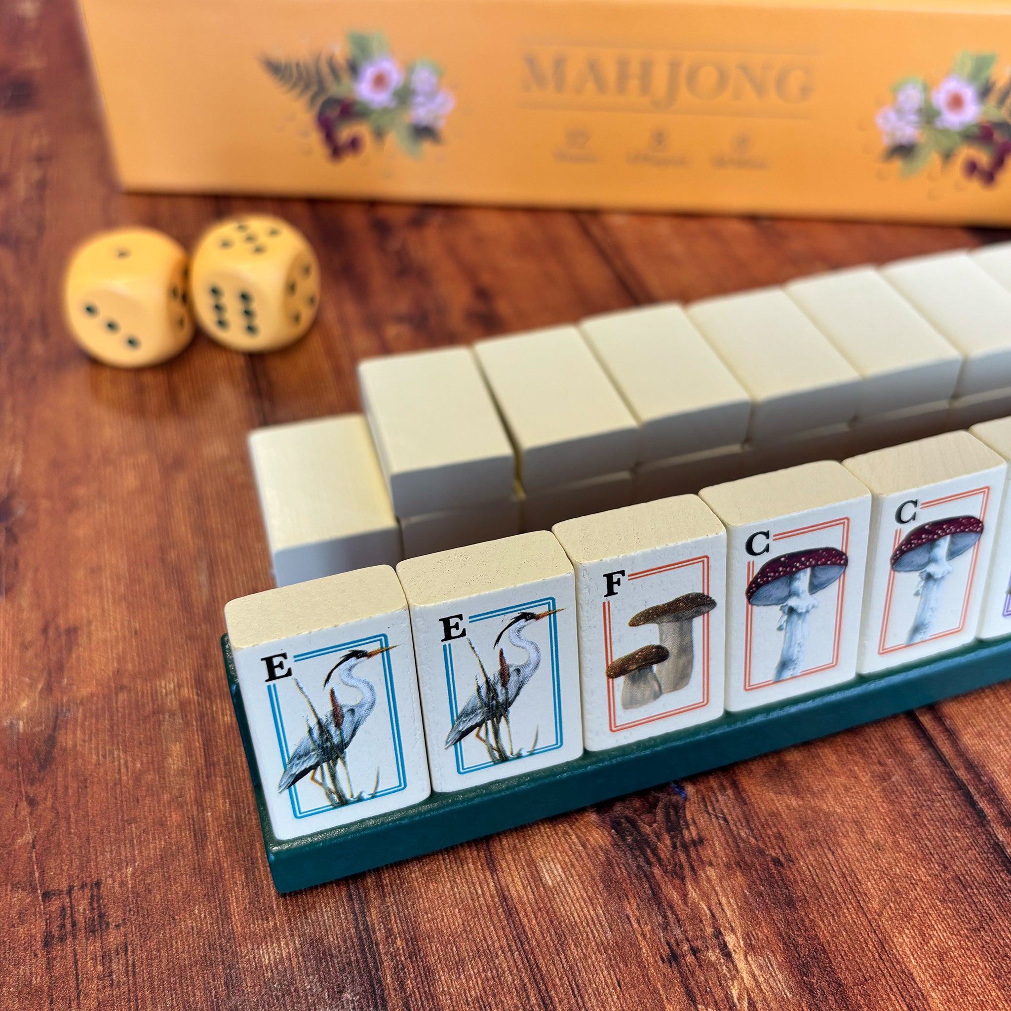 Mahjong game tiles arranged on a wooden table, with two yellow dice and part of the orange Mahjong box visible in the background.