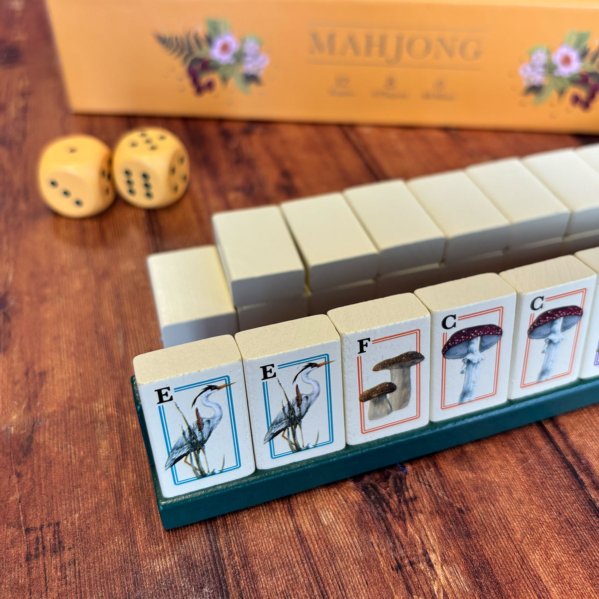 Mahjong game tiles arranged on a wooden table, with two yellow dice and part of the orange Mahjong box visible in the background.
