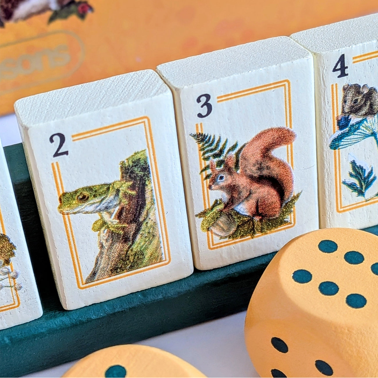Close-up of wooden animal-themed game pieces on a teal board, including numbered cards with illustrated creatures and tan dice in the foreground.