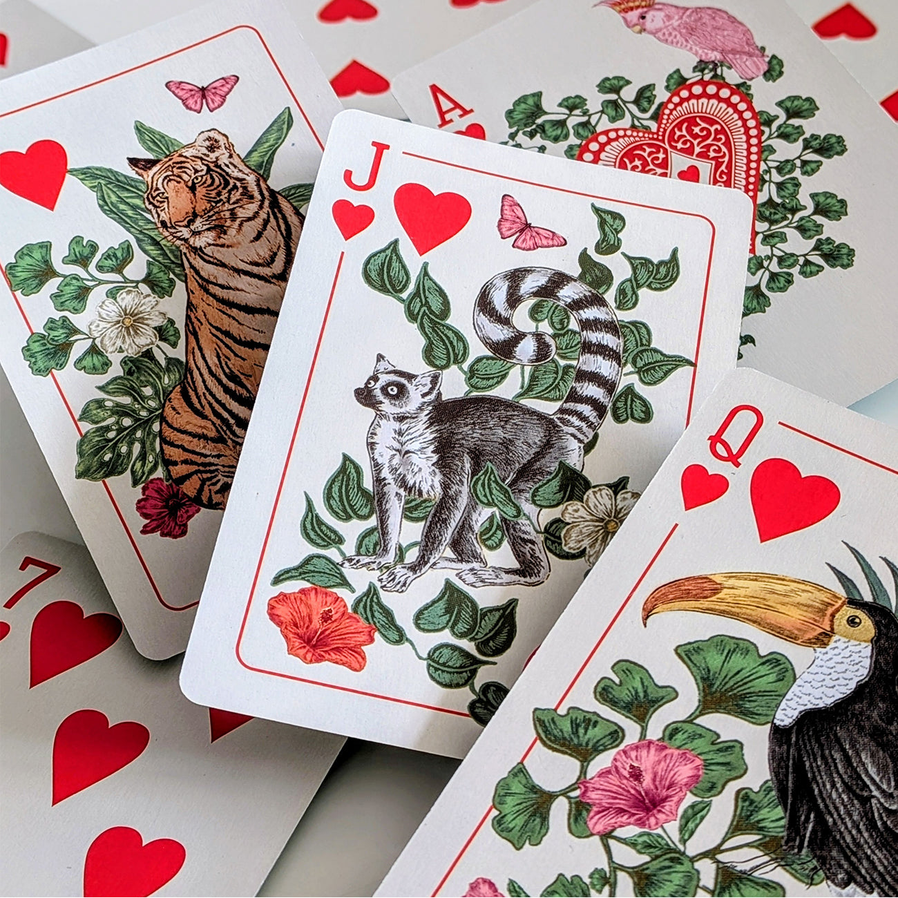 A close-up arrangement of illustrated playing cards featuring animals and floral designs. The central card is a Jack of hearts with a lemur illustration, surrounded by other cards showing a tiger, a queen with a toucan, and red heart suit symbols.
