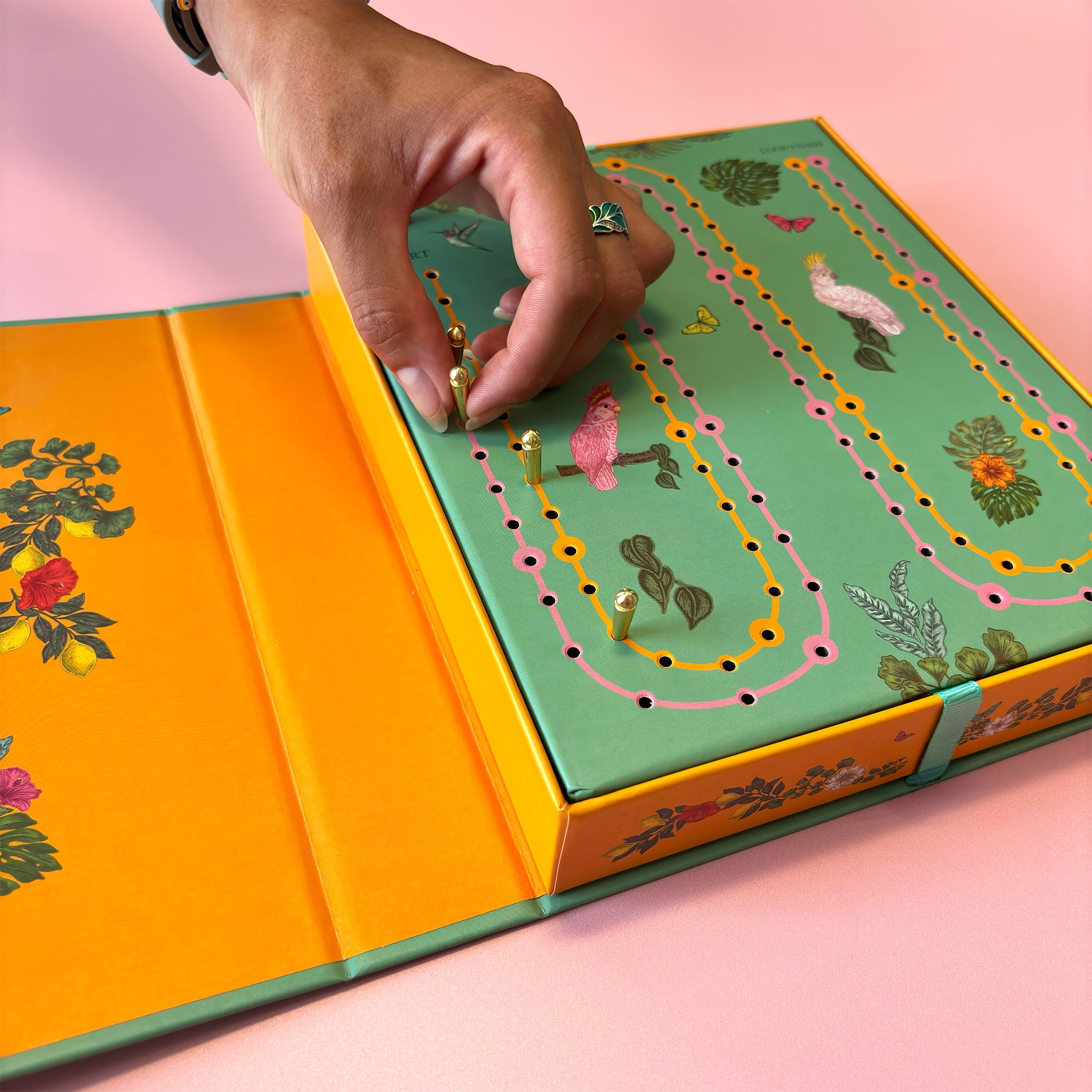 A close-up of a hand placing a small gold bead onto a colourful illustrated board game box. The box has a mint green top with winding pink, yellow, and orange tracks, floral details, and bright orange inner panels, set against a soft pink background.