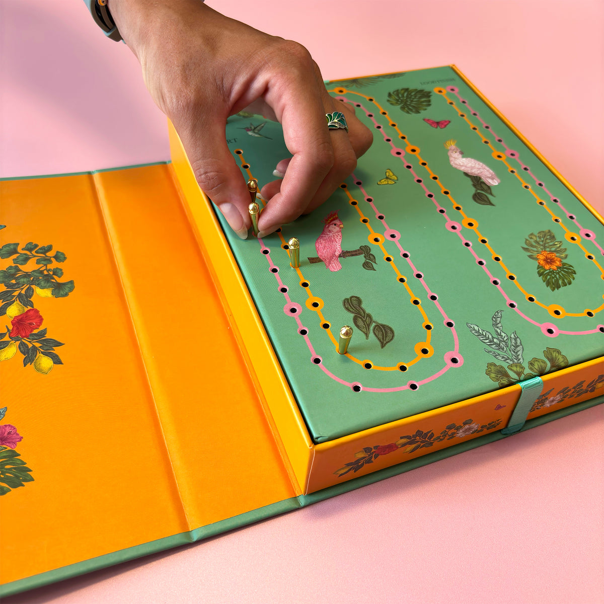 A close-up of a hand placing a small gold bead onto a colourful illustrated board game box. The box has a mint green top with winding pink, yellow, and orange tracks, floral details, and bright orange inner panels, set against a soft pink background.