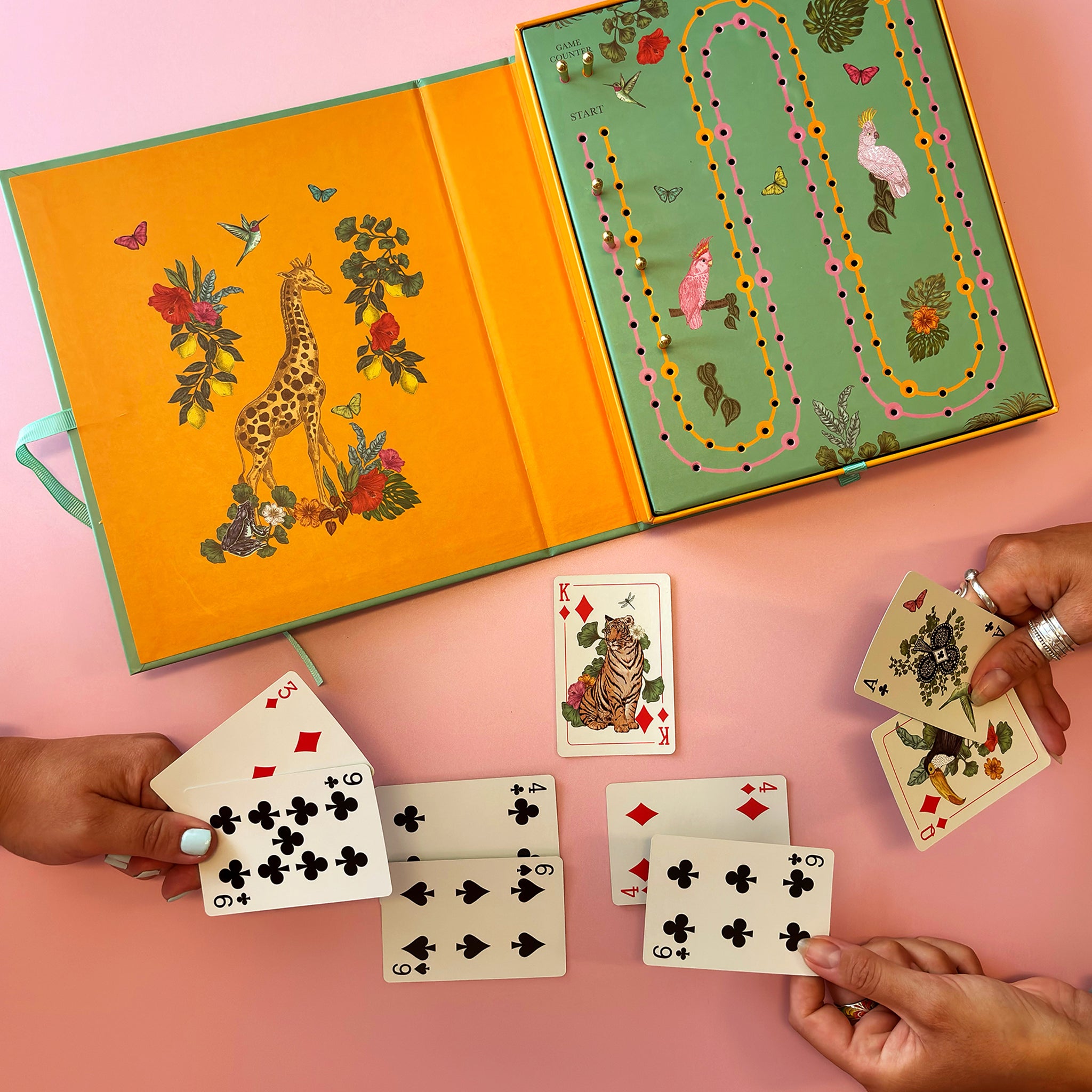A colourful children's card game set displayed on a pink surface, with an open illustrated box showing a giraffe on the cover and a game board on the inside. Several hands are visible holding playing cards, including face-up cards and a deck.