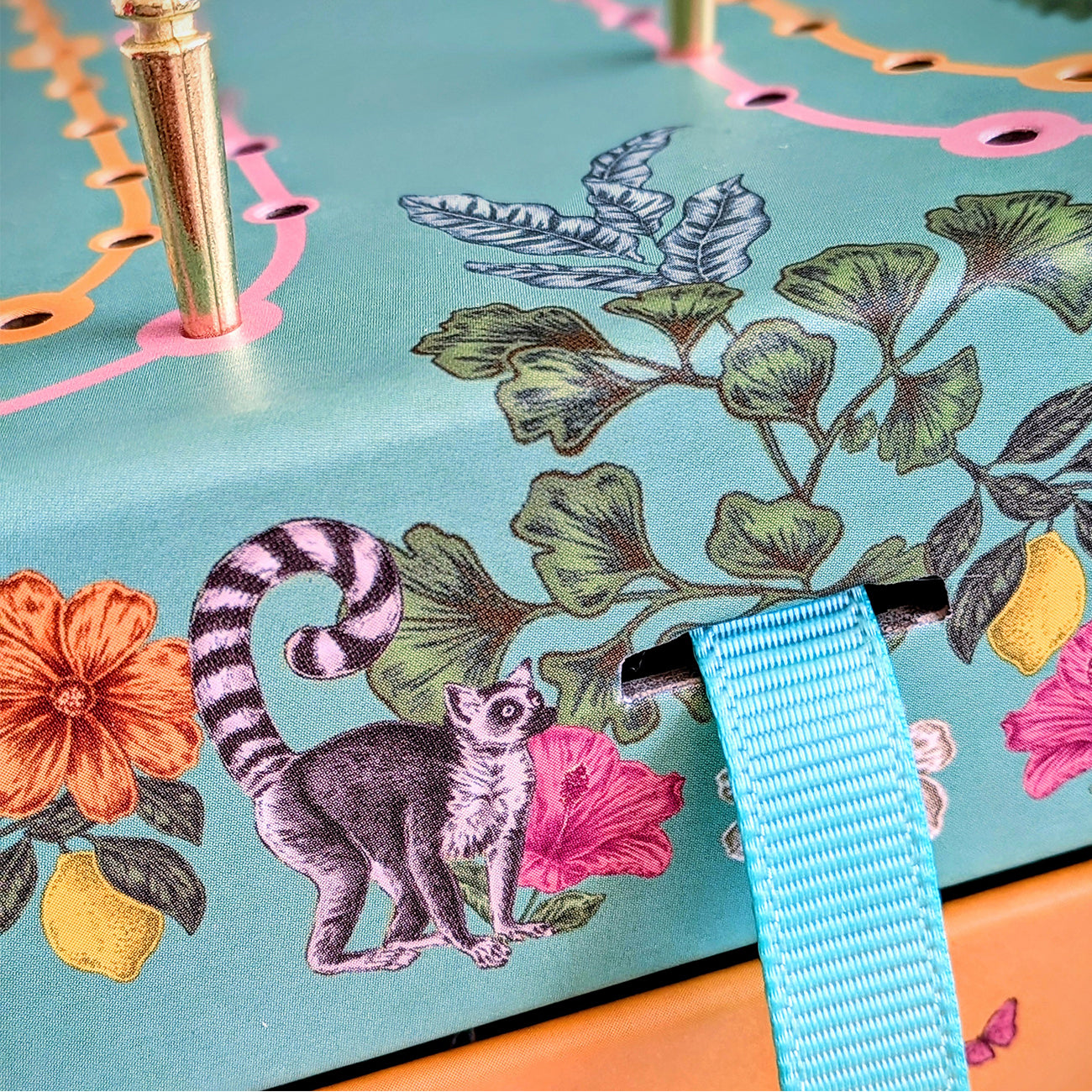 Close-up of a decorative turquoise surface with botanical and animal illustrations, including a lemur, orange flowers, green leaves, and a bright blue ribbon loop.