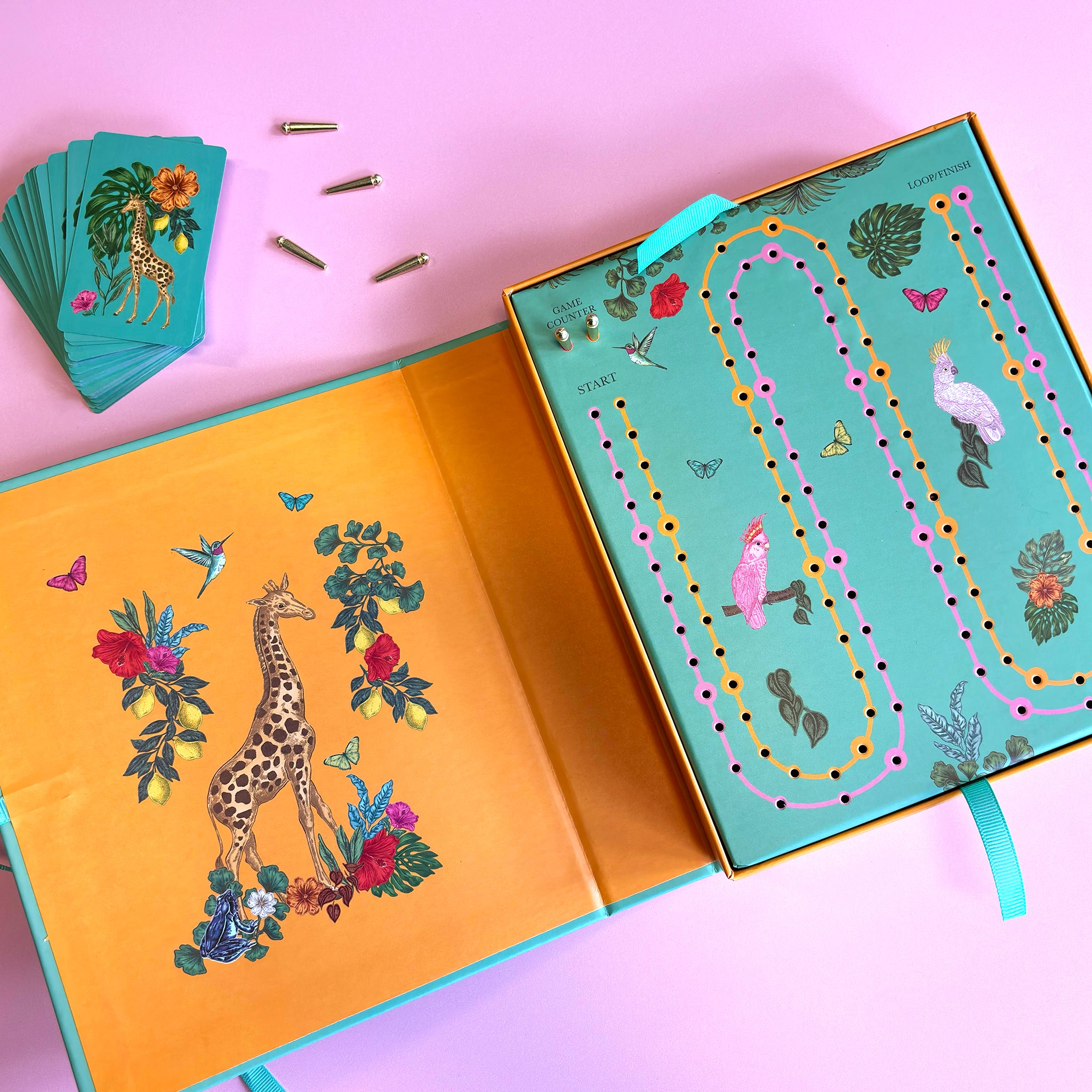 A colourful children's board game set displayed on a pink background, featuring an open game box with illustrated giraffes and butterflies, a matching deck of cards, and several small metal game pieces.