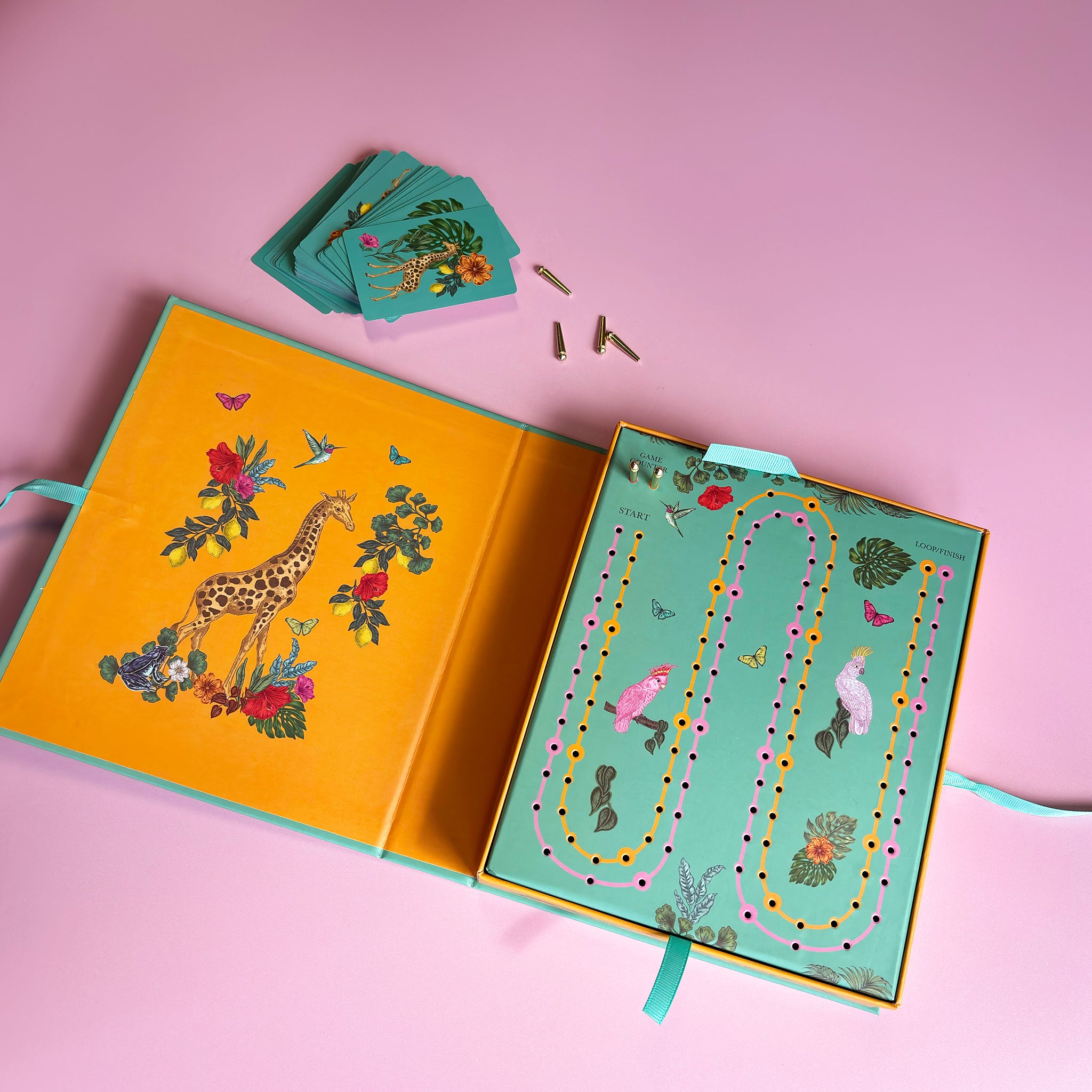 A colourful children's board game set displayed open on a pink background, showing a yellow illustrated cover with a giraffe, a mint green game board with winding path and bird motifs, and several small game pieces.