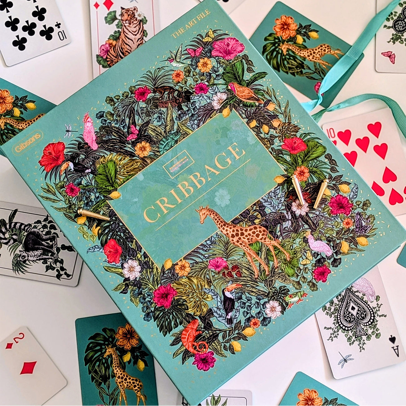 The Artful Cribbage game box displayed in a styled flat-lay arrangement with playing cards and a teal ribbon on a white surface.