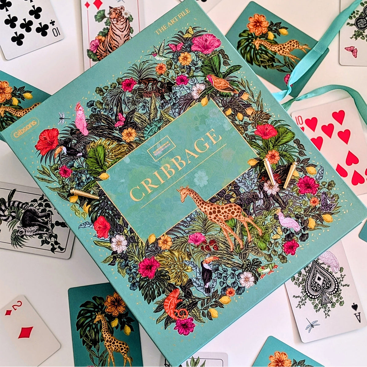 The Artful Cribbage game box displayed in a styled flat-lay arrangement with playing cards and a teal ribbon on a white surface.