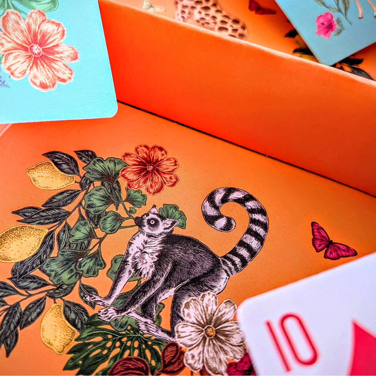 Close-up of an illustrated orange box or card deck interior with a lemur, flowers, leaves, lemons, and butterflies, plus a visible '10' card edge in the foreground.