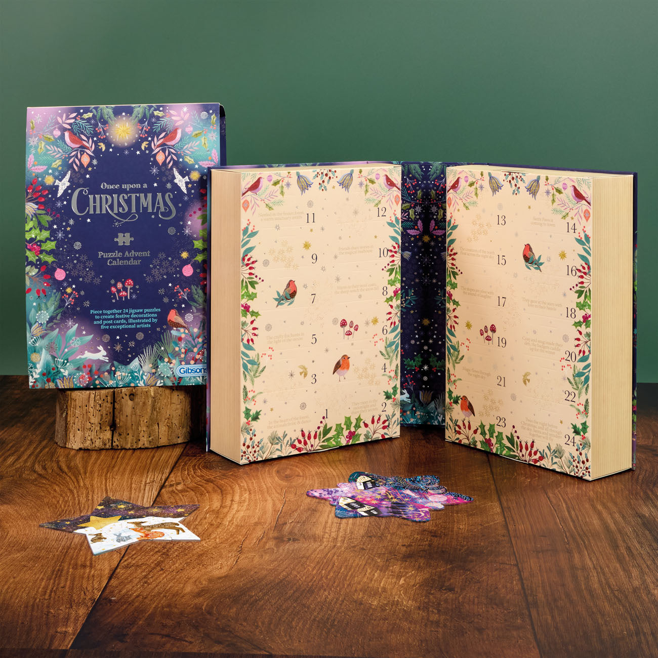 A Christmas advent calendar set displayed on a wooden table against a dark green wall. The set includes a decorative box labelled 'Your evening a Christmas Advent Calendar' on a small wood block, two open illustrated books showing numbered doors, and several colourful puzzle pieces scattered in front.