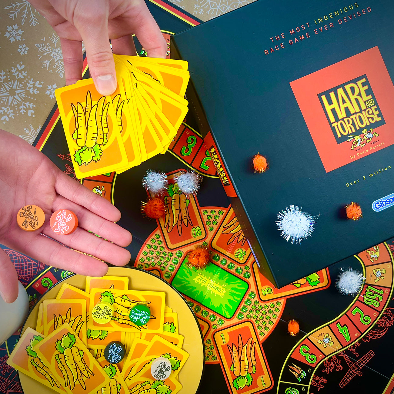 A top-down view of the board game Hare and Tortoise, showing hands holding yellow vegetable cards, a plate of cards, several coloured tokens, and the black game box on a patterned tablecloth.