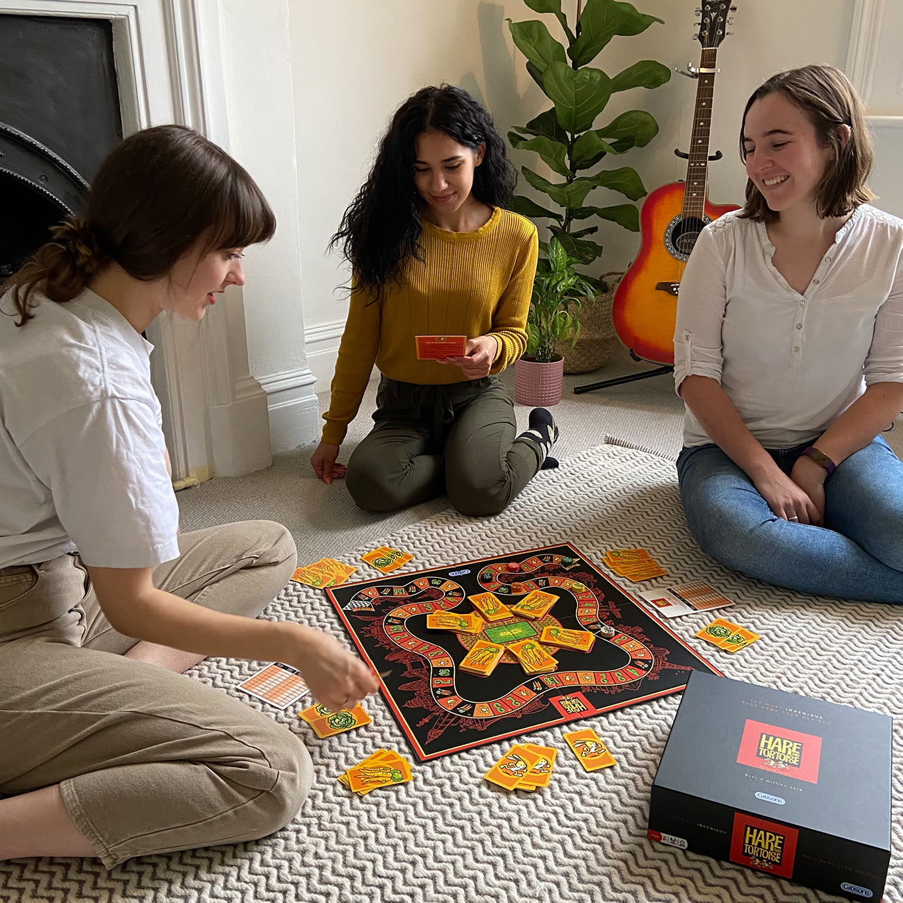Three people sitting on a patterned rug playing a board game indoors. The game board is black with orange and yellow sections, and a black game box labelled 'Hare and Tortoise' sits nearby.