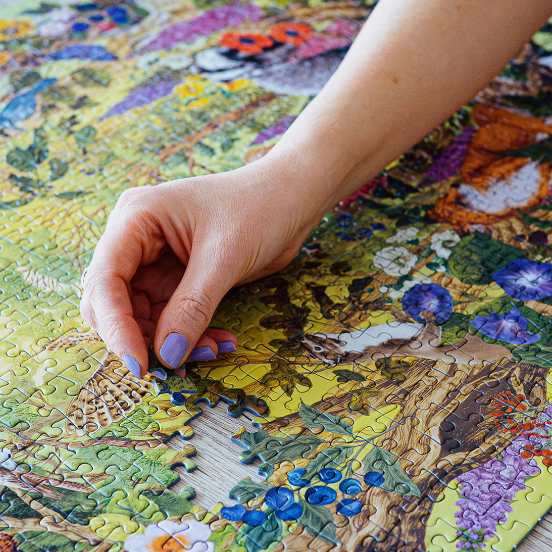Hand with purple nail polish placing a puzzle piece on a colorful puzzle with nature-themed design.