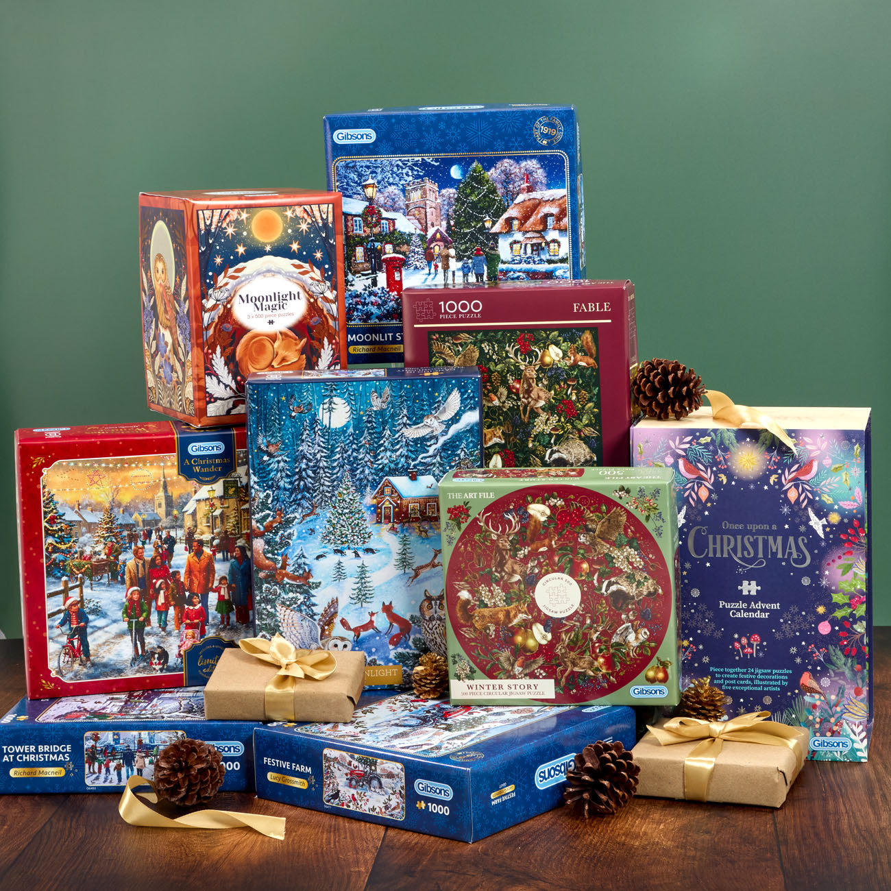 A collection of Christmas-themed jigsaw puzzles in colorful retail boxes, arranged on a wooden surface against a dark green background. Several boxes are wrapped in brown paper with gold ribbons and decorated with pinecones.