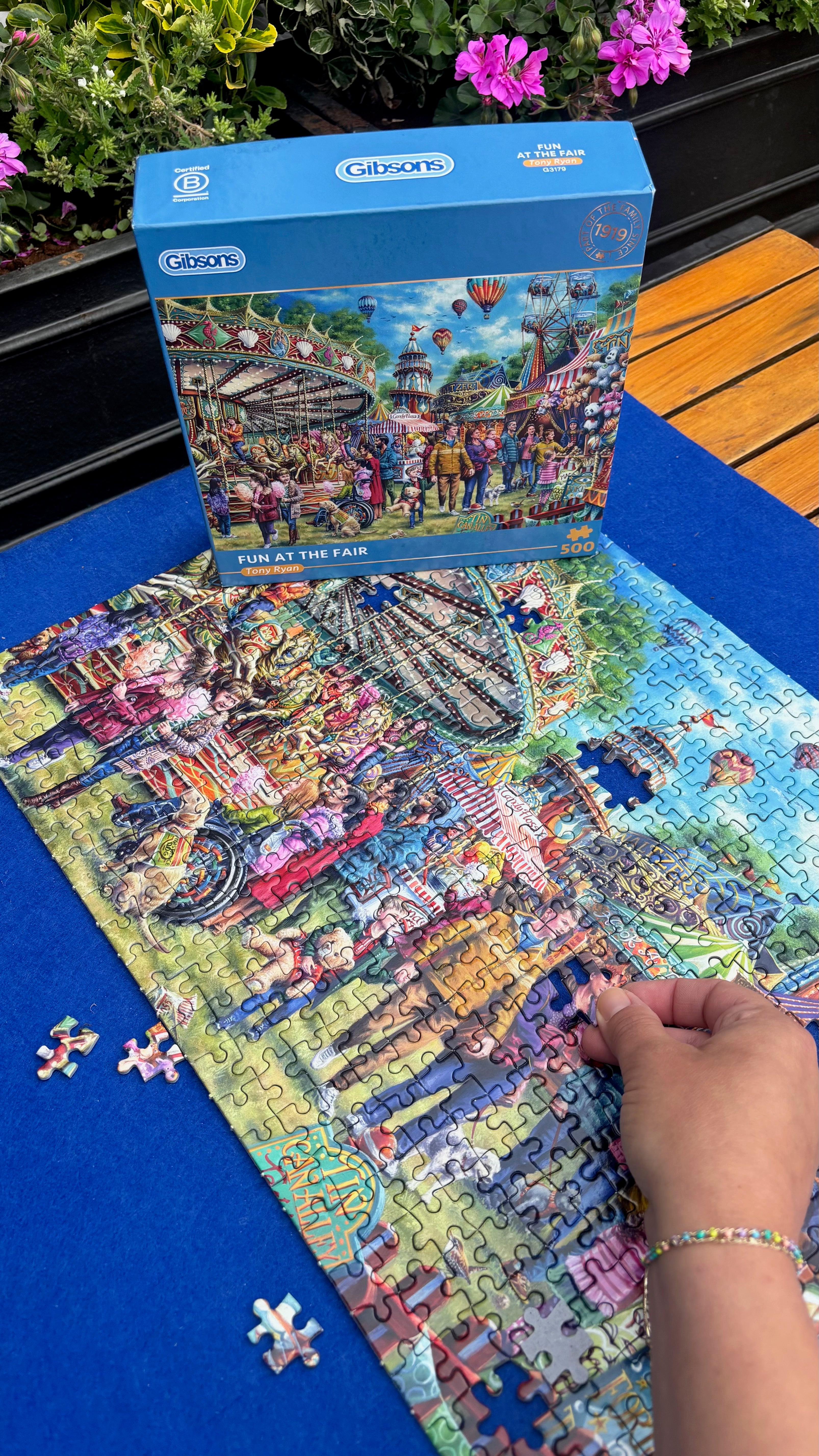 A partially assembled jigsaw puzzle and its blue box are shown on a blue surface outdoors, with a person's hand placing puzzle pieces. The box reads 'Puzzle the Fair' and shows the brand 'Gibsons'.