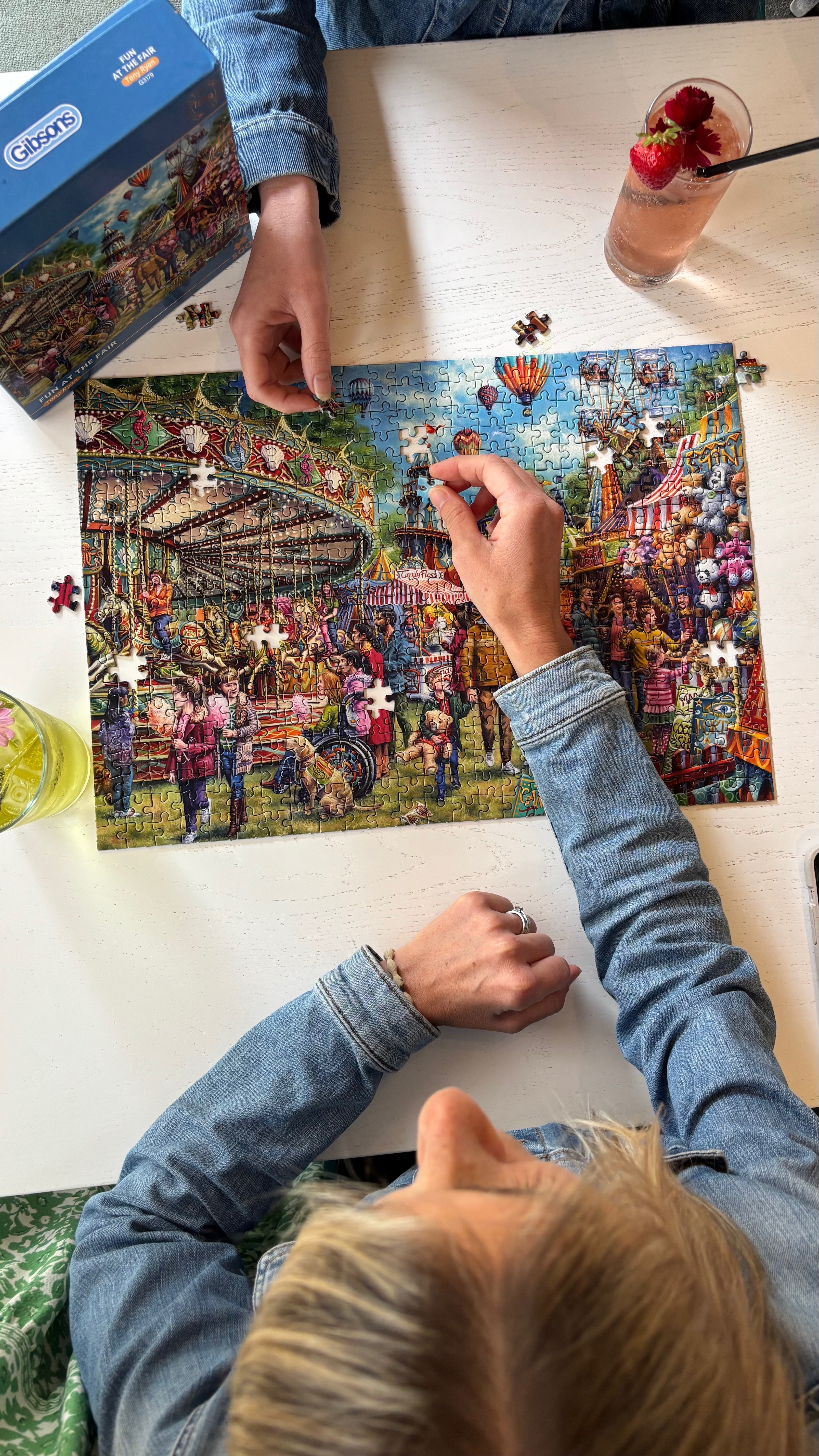 A person placing puzzle pieces on a colorful jigsaw puzzle of a crowded fairground, with the blue jigsaw puzzle box visible in the top left corner on a white tabletop.