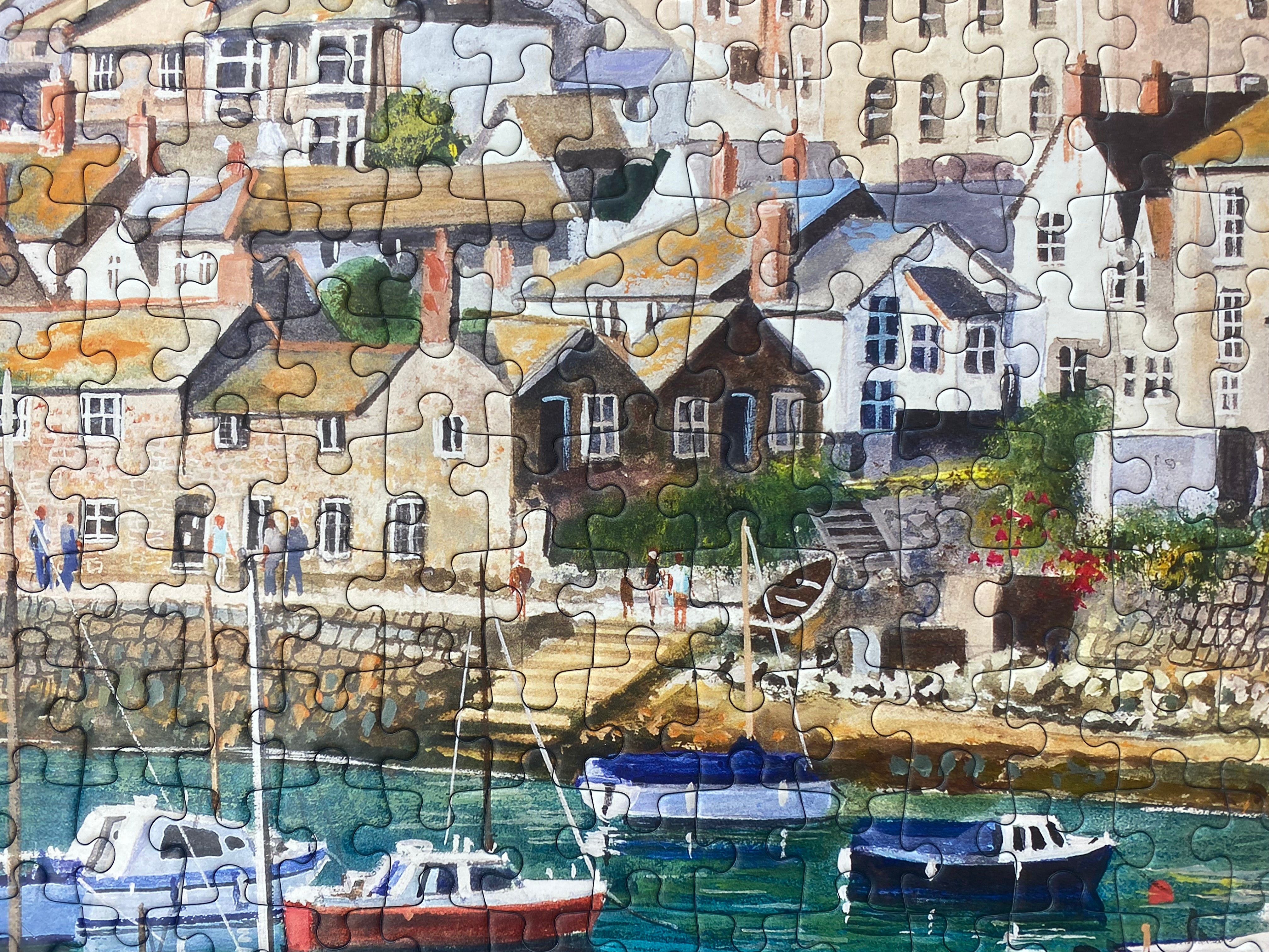 A completed jigsaw puzzle showing a painted coastal village harbor scene with houses, boats, and water in soft pastel and earthy tones.