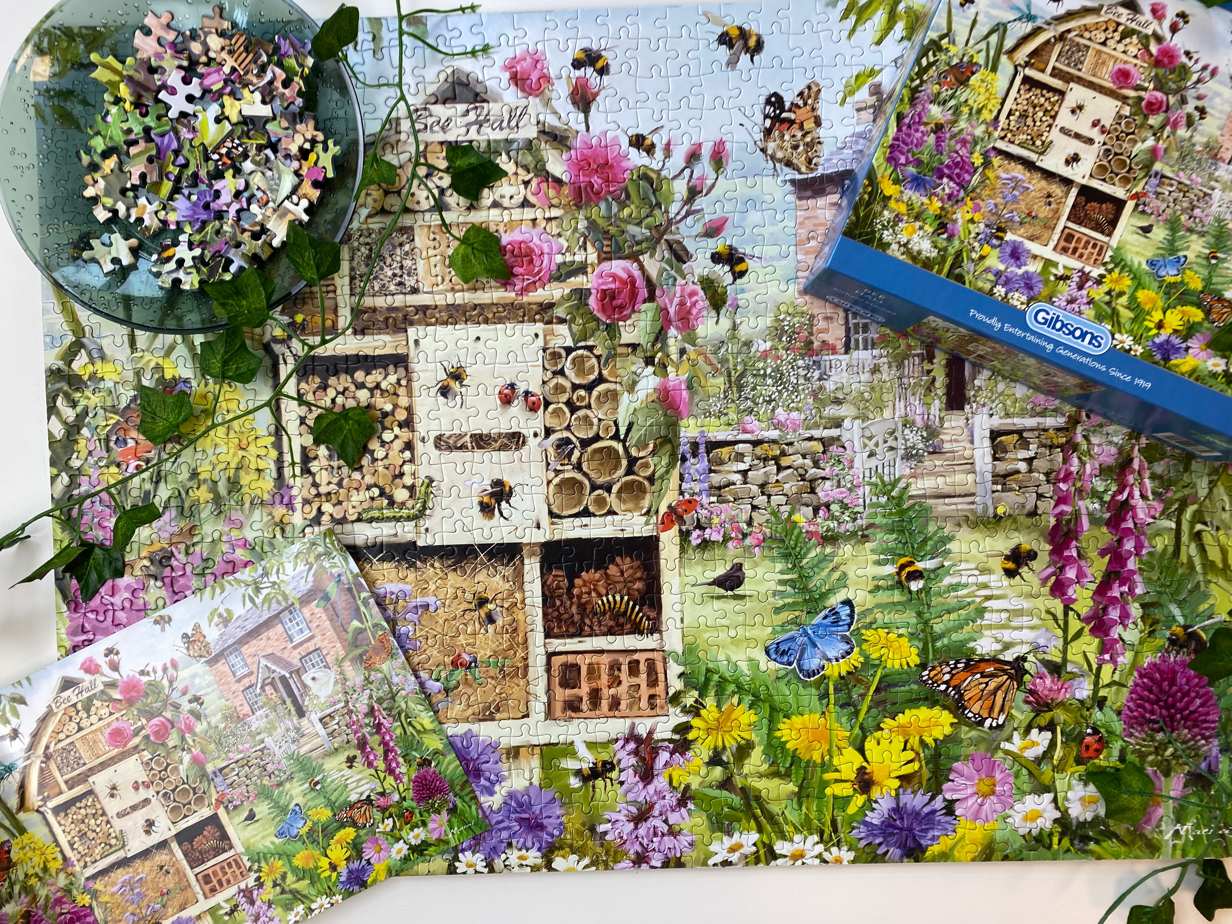 A completed jigsaw puzzle and its box are displayed on a white surface, featuring a colourful garden scene with flowers, bees, butterflies, and a bee house. A small bowl of loose puzzle pieces sits in the upper left corner.