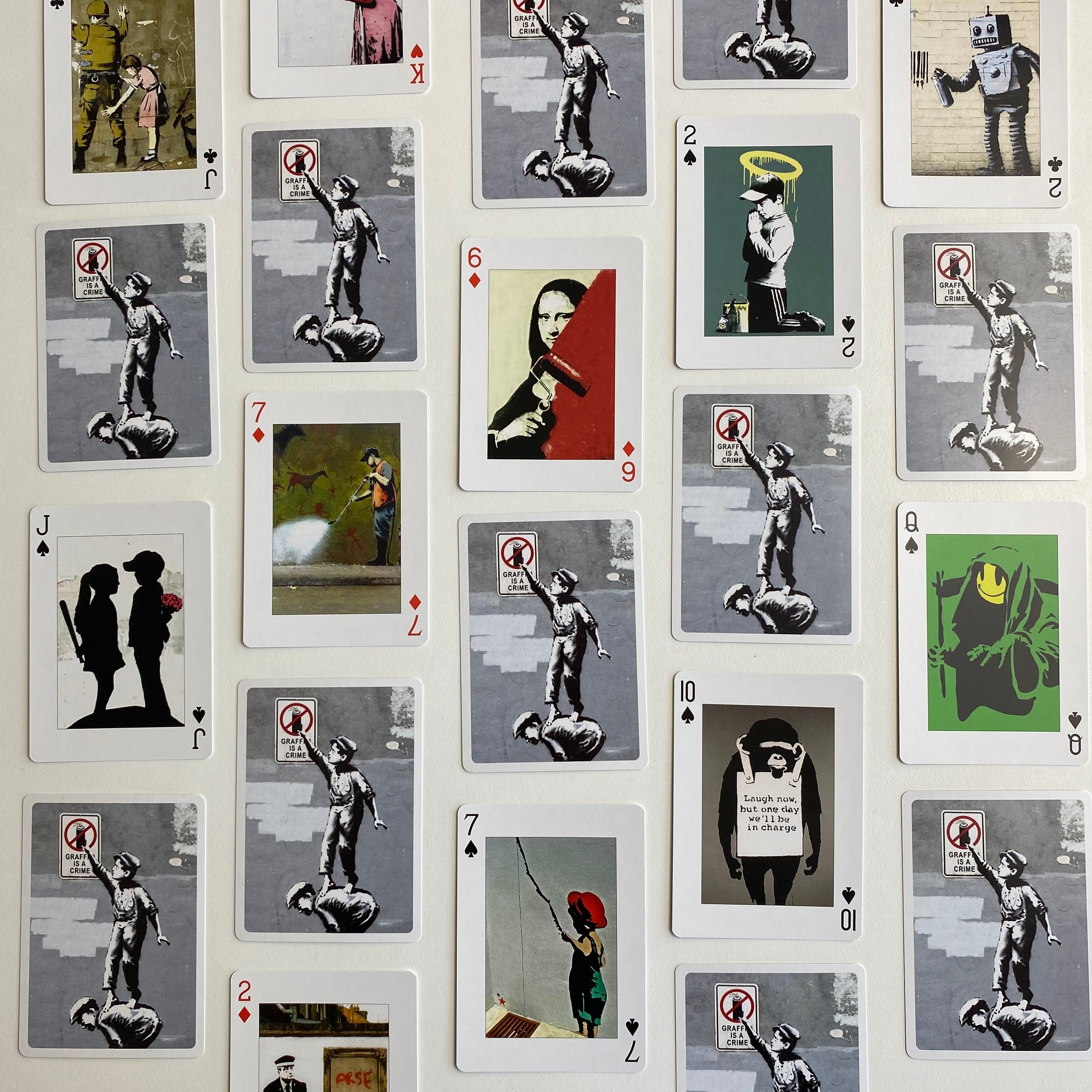 A flat-lay grid arrangement of Banksy-style playing cards featuring street art illustrations, including the Mona Lisa, a robot, a figure with a halo, and silhouettes, on a white background.