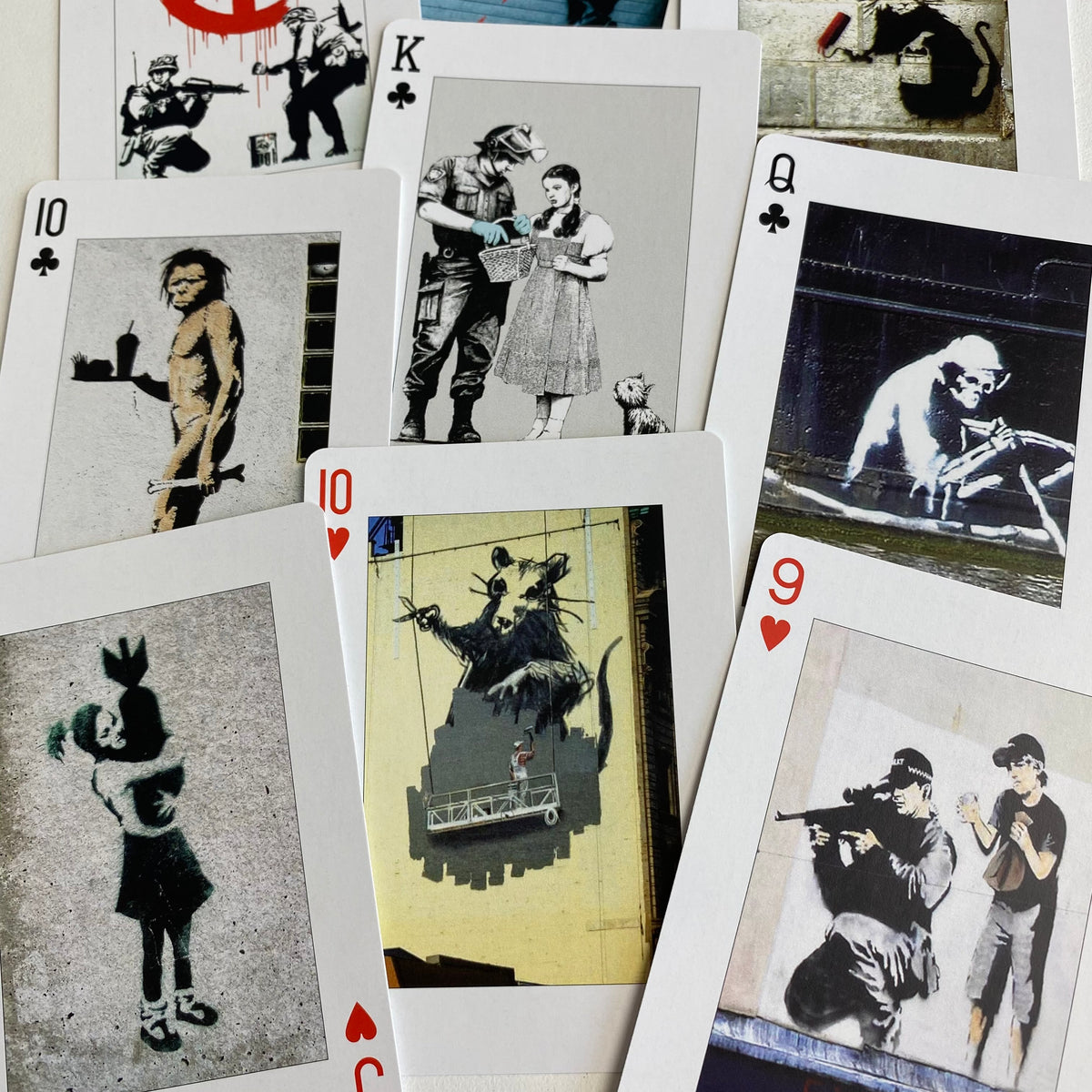 A fan arrangement of playing cards featuring Banksy street art illustrations, including a 10 of clubs, a king of clubs, a queen of clubs, a 10 of clubs, a 10 of hearts, a 9 of hearts, and a 9 of clubs.