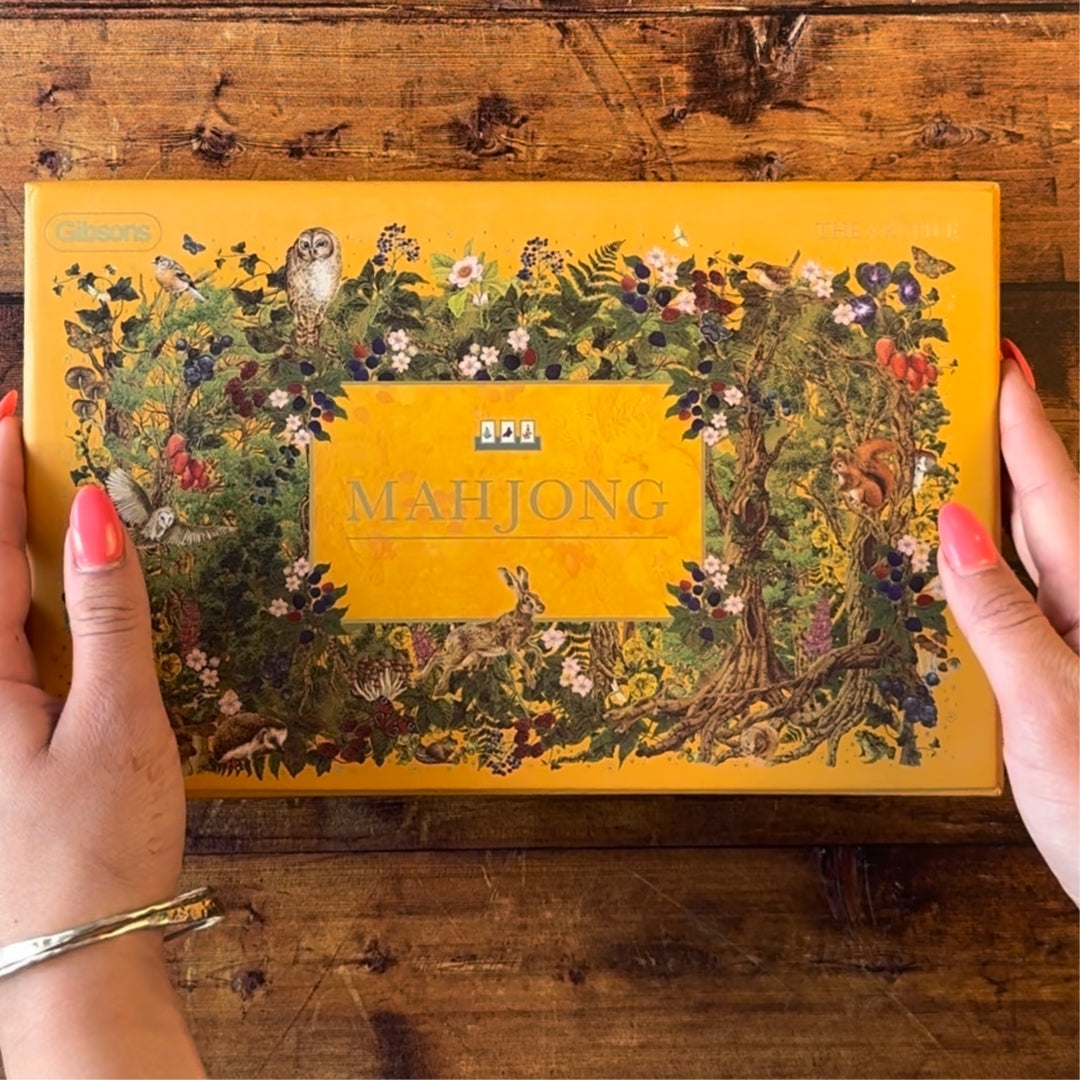 A person's hands with pink nail polish hold a rectangular orange box of ' Mahjong' on a rustic wooden table. The box features an intricate illustration of nature with an owl, rabbit, squirrel, and various flowers and foliage.