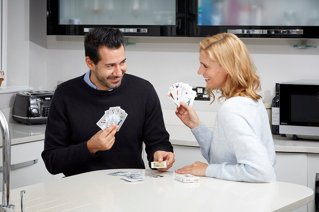 A man and a woman standing at a kitchen counter, smiling and playing with playing cards. The man wears a black sweater over a light blue collared shirt, and the woman wears a light blue sweater.