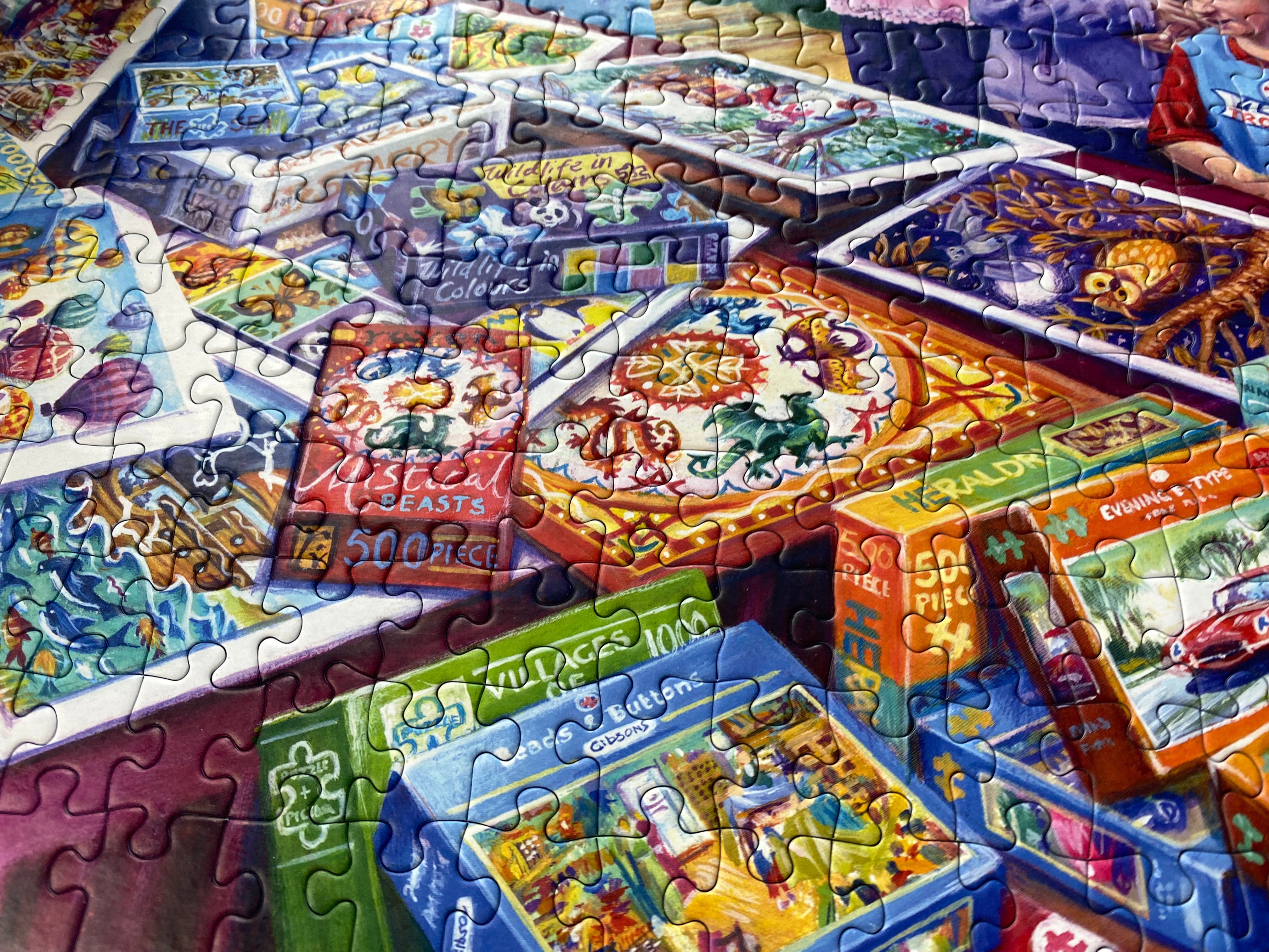 A close-up, angled view of a colourful jigsaw puzzle spread out, with multiple puzzle boxes and completed puzzle pieces visible. The artwork on the puzzle and boxes features bright, whimsical illustrations including a lion, a red car, a green dinosaur, and other playful scenes.