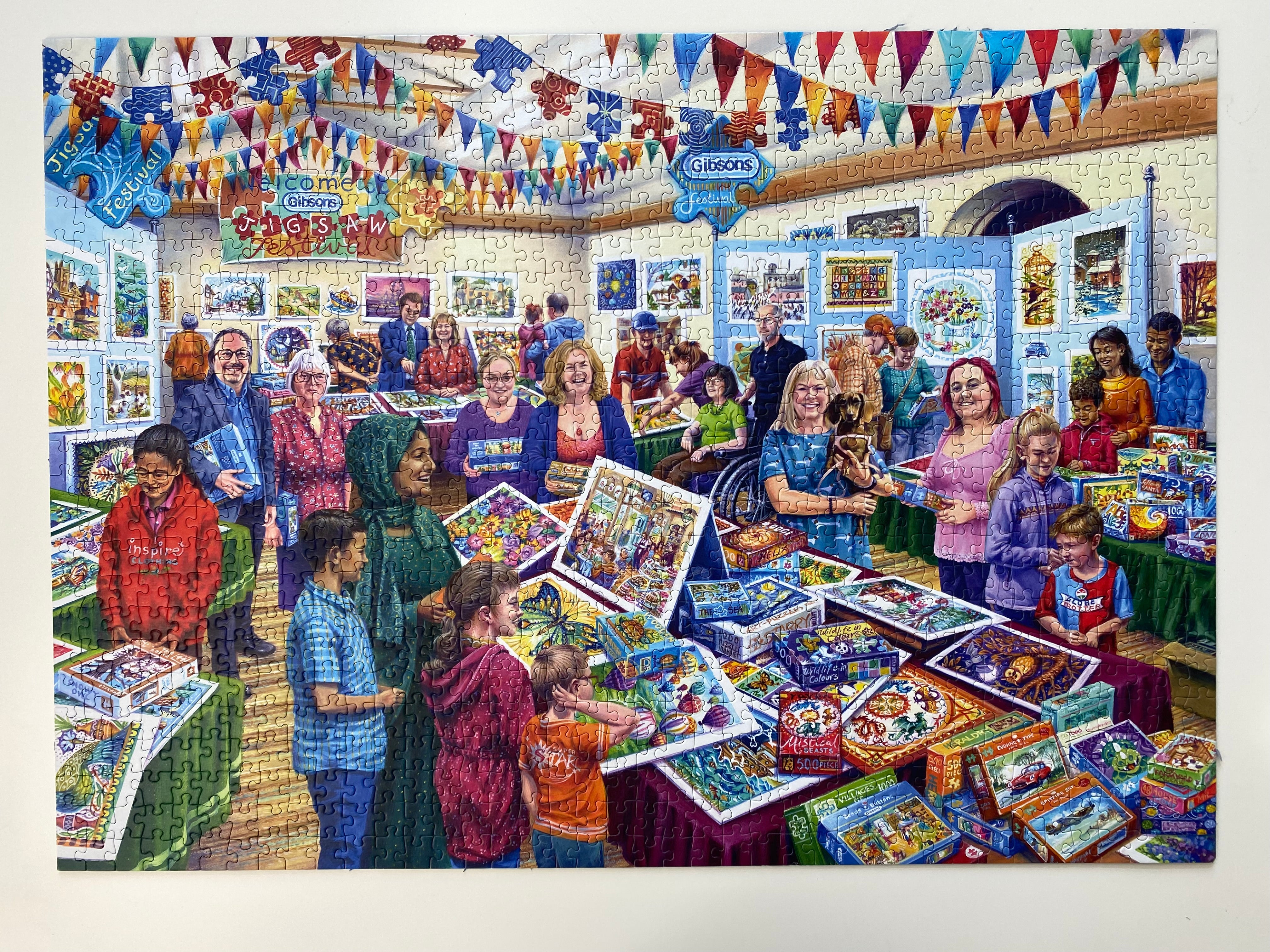 A completed jigsaw puzzle showing a colourful indoor children's book fair or puzzle show, with many people gathered around tables displaying puzzle boxes and artwork. The puzzle is photographed against a plain white background.