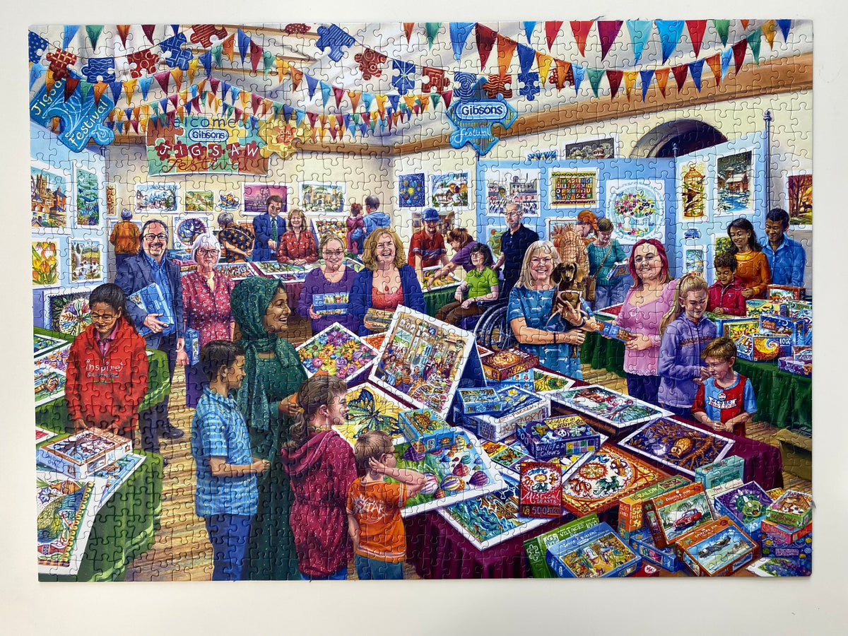A completed jigsaw puzzle showing a colourful indoor children's book fair or puzzle show, with many people gathered around tables displaying puzzle boxes and artwork. The puzzle is photographed against a plain white background.