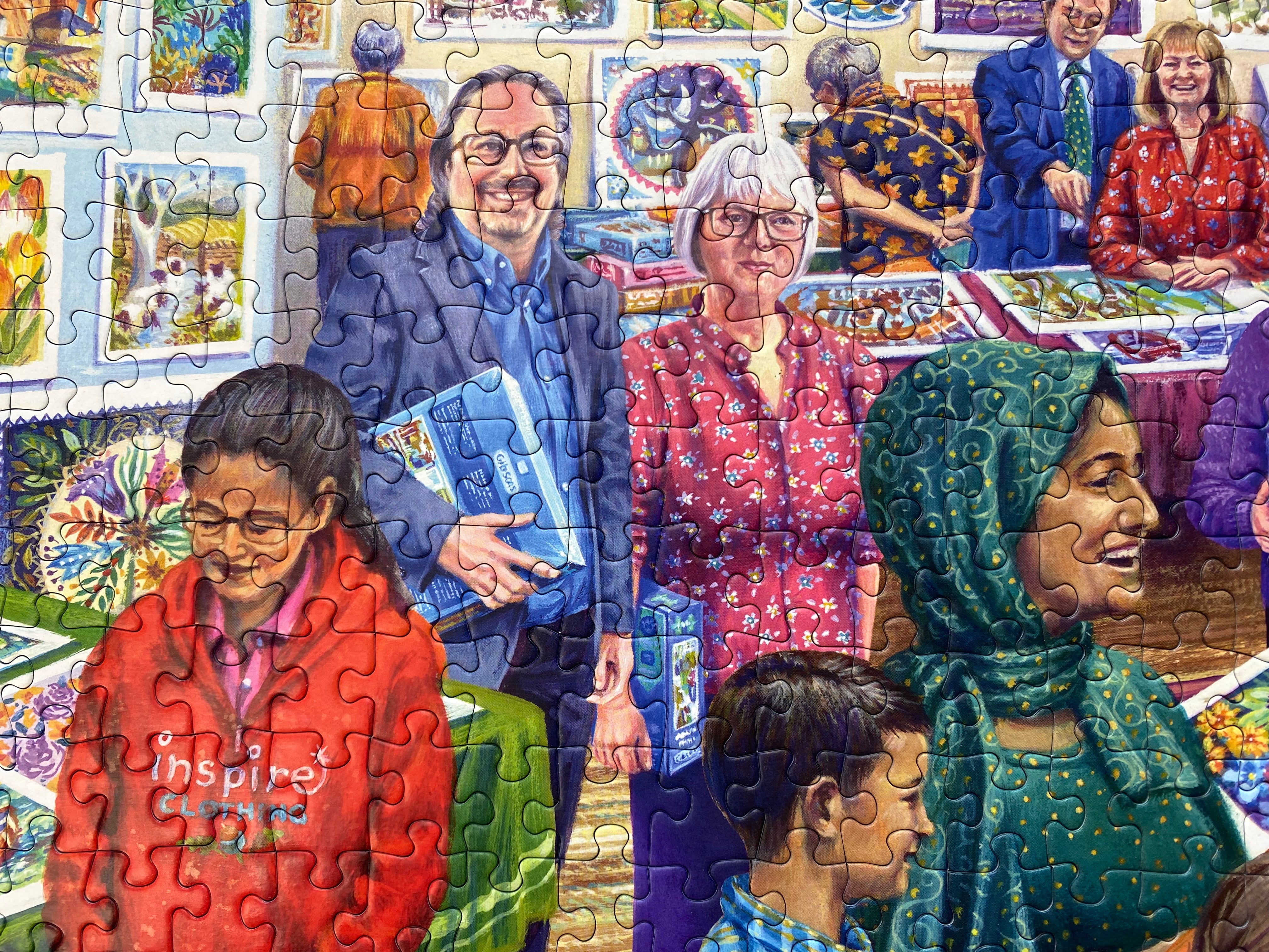 A colourful jigsaw puzzle showing a lively art gallery or exhibition scene with people holding puzzle boxes, surrounded by framed artwork on a wall.