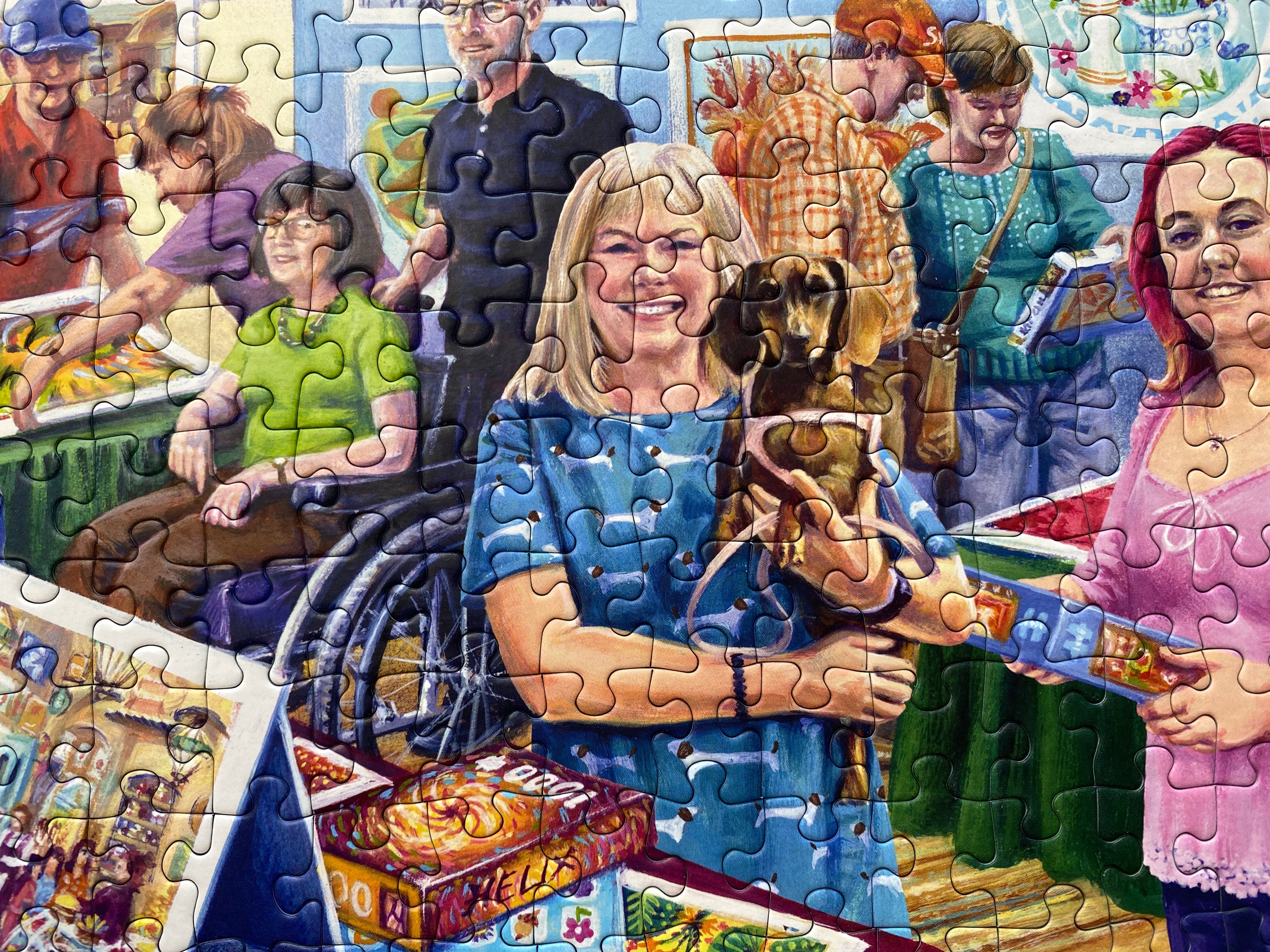 A completed jigsaw puzzle showing a colourful painted scene of people at what appears to be a fair or market, with a smiling woman in the center holding a dog and puzzle pieces forming the image across the whole frame.