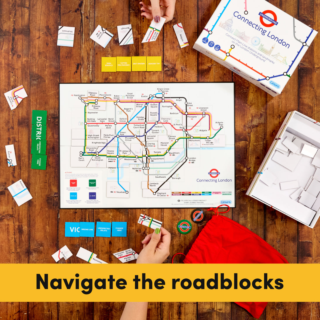 Overhead view of a board game setup on a wooden table, featuring a Connecting London transit map board, game pieces, cards, a red drawstring bag, and open packaging. A yellow banner at the bottom reads 'Navigate the roadblocks'.