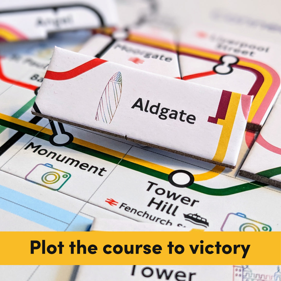 A close-up of a board game featuring a white card labeled 'Aldgate' with a striped sailboat icon, placed over a colourful transit-map style board with station names like Tower Hill and Monument. A yellow banner at the bottom reads 'Plot the course to victory'.