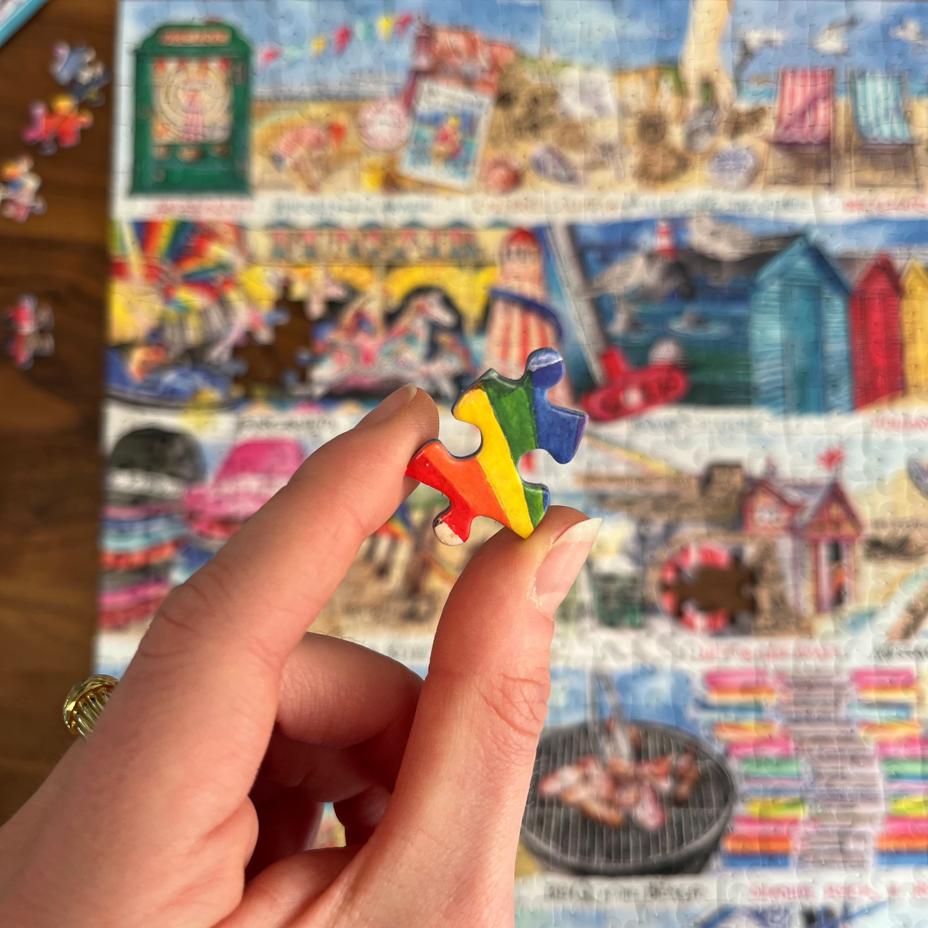 A hand holds a small, colorful jigsaw puzzle piece in front of a large, detailed multicolor puzzle that is partially assembled on a wooden surface.