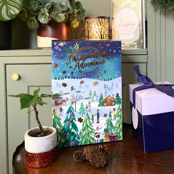 A Christmas advent calendar next to a small holly tree and a bow-wrapped present on a brown table.