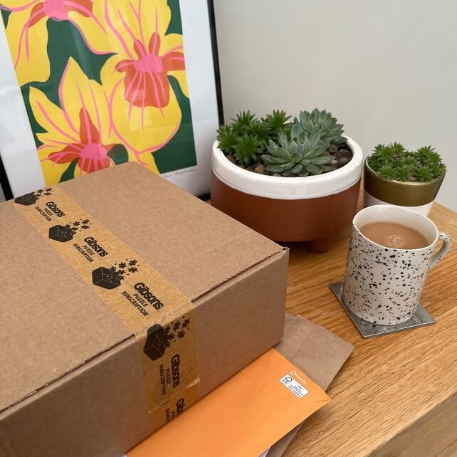 Gibsons puzzle subscription package delivery on a wooden table with a hot beverage.